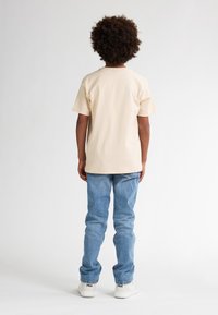 Beige katoenen t-shirt met korte mouwen en een relaxte pasvorm, gecombineerd met lichtblauwe jeans en witte sneakers. Gezien vanaf de achterkant.