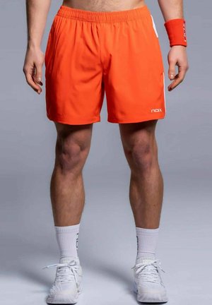 Mann trägt leuchtend orangefarbene Sportshorts, weiße Sportschuhe und weiße Socken, steht vor grauem Hintergrund.
