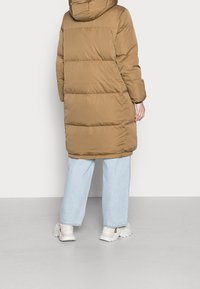 Lungo cappotto voluminoso di colore marrone, con cappuccio e dettagli delle cuciture. Indossato sopra jeans azzurri chiari e sneakers bianche spesse.