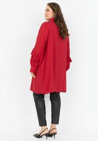 Sieviete stāv un skatās sānis, valkājot garu sarkanu blūzi ar volāniem, melnu ādas bikses un melnus kaķu papēžu apavus.