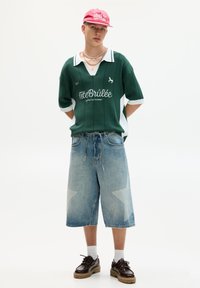 Grøn, krave rugby skjorte med hvide detaljer; brede, falmede denim shorts med stjernesymbolet; mørkebrune sko; lyserød kasket.