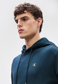 Felpa blu navy in misto cotone, con tasca a marsupio frontale, logo dorato sul petto e cordini regolabili sul cappuccio.