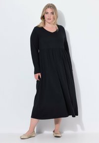 Robe noire à manches longues avec un décolleté en V, jupette évasée et taille froncée. Le modèle l'a assortie avec des chaussures plates beiges. Texture de tissu lisse.