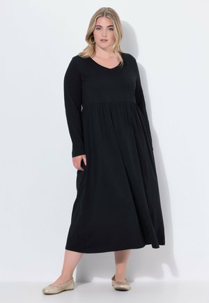 Schwarzes Langarmkleid mit V-Ausschnitt, ausgestelltem Rock und gerafftem Taillenbereich. Das Model kombinierte es mit beigen flachen Schuhen. Glatte Stoffoberfläche.