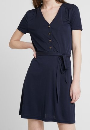 Femme portant une robe bleu marine mi-longue avec des manches courtes, un col en V, des boutons sur le devant et une ceinture nouée à la taille.