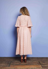 Mulher de pé descalça no chão de madeira, de frente para uma parede azul, vestindo um longo vestido de renda rosa com mangas largas e cabelo encaracolado solto.