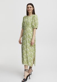Robe midi fleurie verte avec des manches bouffantes, une taille froncée, des fentes sur les côtés et de petites fleurs blanches. Portée avec des sandales à talons hauts noires à lanières.