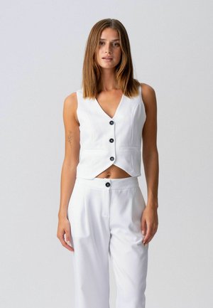 Frau mit glattem, schulterlangem braunem Haar, die eine weiße, knöpfbare ärmellose Weste und passende weiße Hose vor einem grauen Hintergrund trägt.