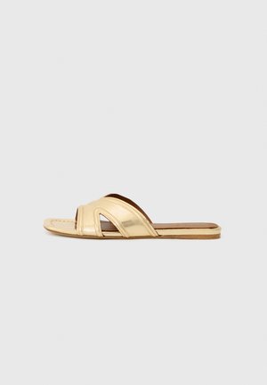 Sandale à talon plat en cuir doré présentant un design à bout ouvert, une forme minimaliste et des accents découpés sur la bride. Semelle plate avec un intérieur marron.