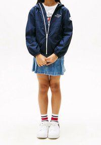 Námořnická větrovka s kapucí a bílým zipem, lehký materiál; nošena přes tričko s potiskem a džínsovou sukni, doplněná bílými teniskami a pruhovanými ponožkami.