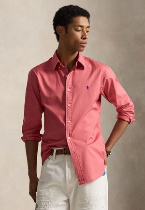 Νέος άνδρας που φοράει κοραλλί πουκάμισο με κουμπιά και σηκωμένα μανίκια και λευκό ξεφτισμένο παντελόνι, στέκεται με το ένα χέρι στη μέση.