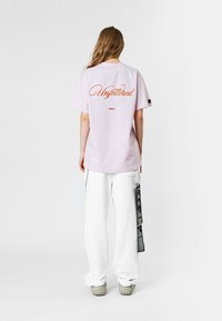 Camiseta de manga corta rosa con "Unfettered" en letras naranjas en la parte posterior, combinada con pantalones holgados blancos y calzado gris.