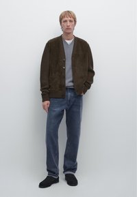 Brauner Wildleder-Cardigan über einem grauen Pullover, blaue Jeans und schwarze Schuhe. Der Cardigan hat gerippte Ärmel und eine durchgehende Knopfleiste.