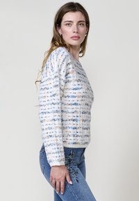 Pull en tricot crème avec des rayures horizontales bleues et orange, motif texturé, col rond et manches longues. Associé à un jean brodé.