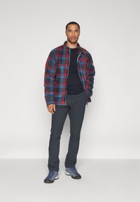 Kockovaný fleece sveter v modrej, červenej a čiernej farbe, skombinovaný s čiernym tričkom a tmavými nohavicami. Obuv je šed�á s modrými šnúrkami, s robustným dizajnom.