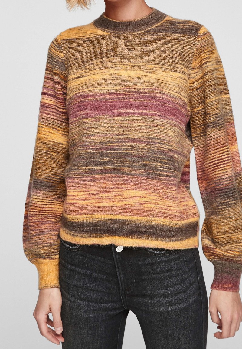 Femme portant un pull tricoté à rayures multicolores avec des manches bouffantes et un jean noir, visible du cou jusqu'aux hanches.