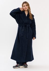 Donna con lunghi capelli che indossa un cappotto peloso blu scuro con cintura e sneakers colorati, si trova con una mano vicino all'orecchio su uno sfondo bianco.