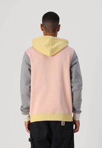 Sudadera con capucha en rosa, gris y amarillo. Presenta un bolsillo tipo canguro, puños acanalados y una textura suave. La vista trasera destaca el uso del color en bloques.
