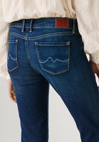 Πίσω όψη ατόμου που φοράει σκούρα μπλε τζιν με πίσω τσέπες που έχουν κυματιστά λευκά ραφτά και δερμάτινο σήμα μάρκας στη ζώνη μέσης.