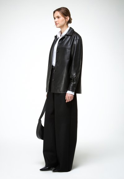 Nő áll fekete bőr gombos dzsekiben, fehér ingben, széles szárú fekete nadrágban, fekete hegyes orrú cipőben, kezében fekete válltáskával.