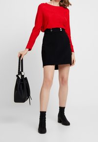 Roter, strukturiert gestrickter Pullover mit langen Ärmeln, kombiniert mit einem schwarzen Minirock mit Knopfverzierungen und Taschen. Schwarze Bucket-Tasche und Stiefeletten.