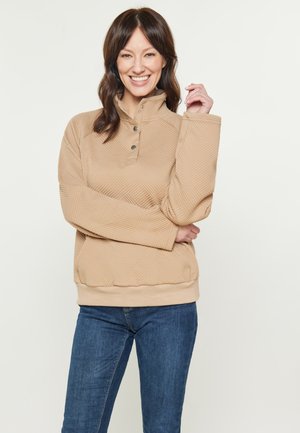 Beige quiltet pullover med høj krave, tre knapper og strukturerede diagonale mønstre; parret med mørkeblå jeans.
