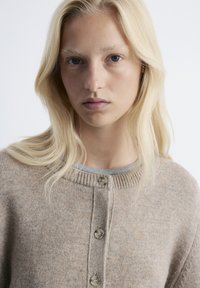 Beiger Strickcardigan mit rundem Halsausschnitt, ausgestattet mit fünf Knöpfen und einem gerippten Muster, über einem grauen Oberteil getragen. Die Haare sind lang und glatt.