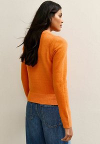 Femme aux longs cheveux bruns, portant un pull orange et un jean bleu, se tenant de profil contre un mur beige uni.