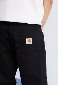 Pantalones de mezclilla negros con un corte recto, con una etiqueta blanca que tiene un logotipo naranja en el bolsillo trasero derecho. Textura suave y diseño limpio.