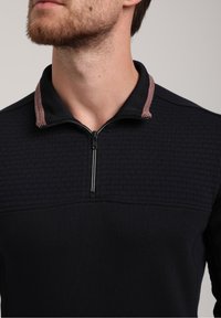 Schwarzer Pullover mit einem strukturierten oberen Bereich und einem Reißverschlusskragen. Der Kragen hat ein kontrastierendes rot-weißes Streifenmuster. Weiches Material.
