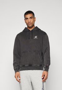Haut à capuche gris foncé en coton, avec une poche kangourou, un capuchon à cordon ajustable, et un logo blanc brodé sur la poitrine.