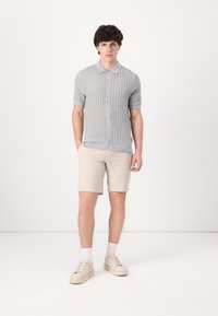 Cardigan gri tricotat cu mânecă scurtă, încheiat cu nasturi, asortat cu pantaloni scurți bej deschis și adidași bej. Design simplu și texturat.