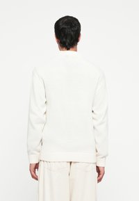 Pulover alb din tricot cu un tiv și mansete cu nervuri, având un guler înalt. Țesătura are o suprafață texturată și un design simplu, minimalist.