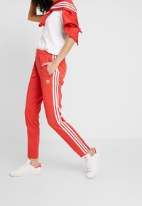 Červené tepláky se bílými bočními pruhy a logem Adidas. Kombinované s bílým tričkem a bílými teniskami s oranžovými akcenty.