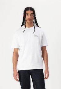 Hvit bomulls T-skjorte med rund hals og korte ermer, med en subtil grå logo på venstre bryst. Bært sammen med mørk denim jeans.