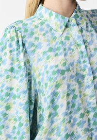 Leichte, mehrfarbige Bluse mit Kragen. Sie hat ein blau-grünes, abstraktes Muster, einen strukturierten Stoff und lange Ärmel.