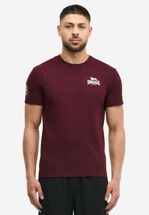 Homme portant un t-shirt bordeaux ajusté de Lonsdale London et un pantalon noir à cordon, debout devant un fond blanc uni.