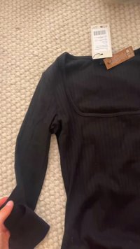 Zwarte langemouwen top van ribstof, met een vierkante halslijn en een label aan de kraag, gepresenteerd op een gestructureerd oppervlak.