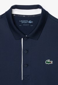 Marinblå pikétröja med en två-knapplist, vit detalj på kragen och en grön krokodil-logotyp på vänster bröst. Normal passform.