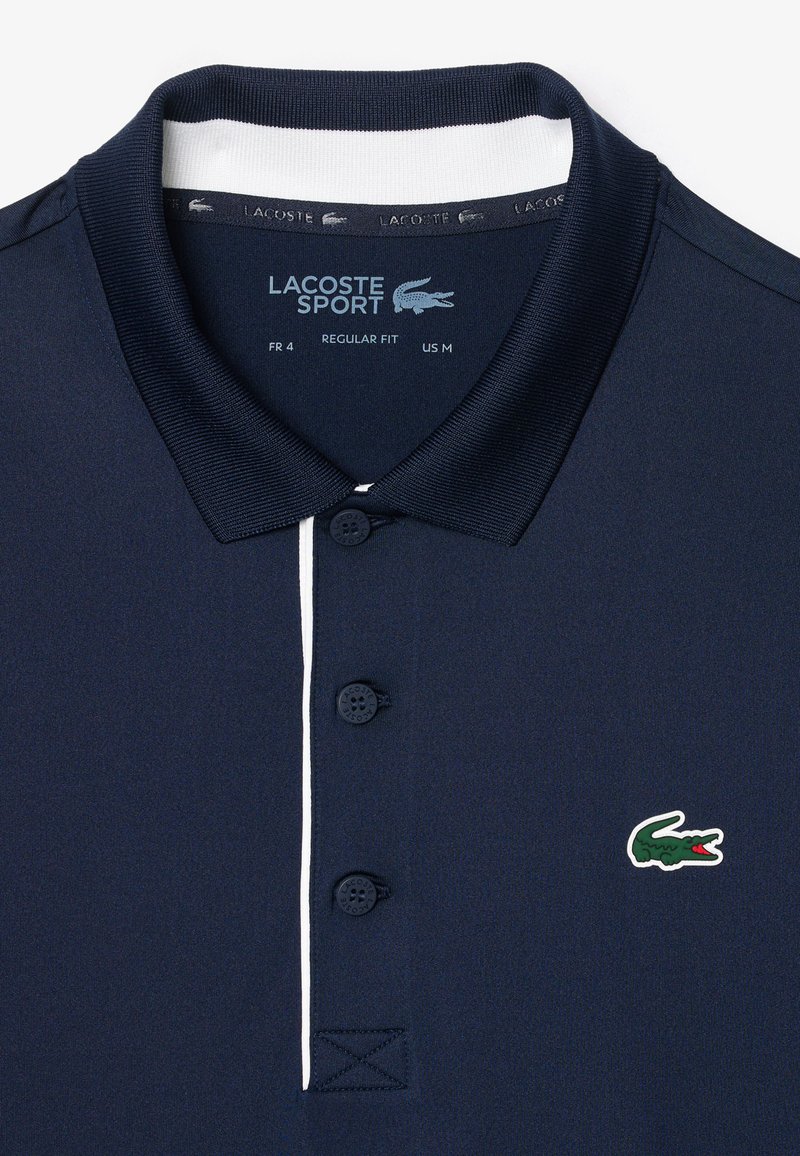 Polo shirt blu navy con apertura a due bottoni, dettaglio bianco sul colletto e logo di coccodrillo verde sul lato sinistro del petto. Vestibilità regolare.