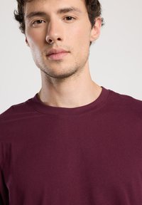 Young man with light skin and short curly brown hair wearing a plain dark maroon crew-neck shirt, facing forward.