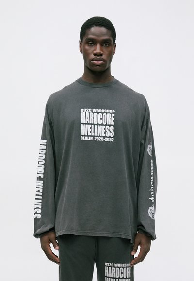 Mladý muž v tmavošedom tričku s dlhým rukávom a nohaviciach, na ktorých je biele písmo "Hardcore Wellness" a názov mesta "Berlín 2025-2032."