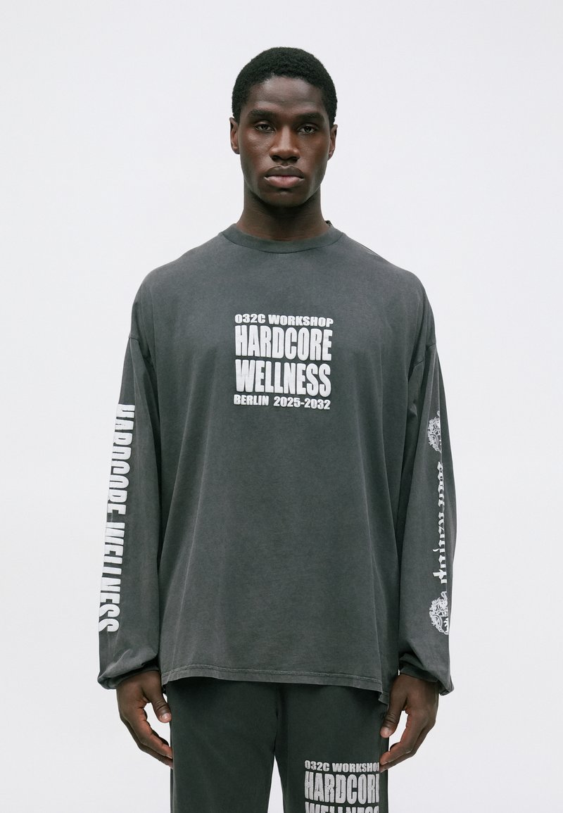 Junger Mann in einem dunkelgrauen Langarmshirt und Hose mit weißem Text "Hardcore Wellness" und dem Stadtnamen "Berlin 2025-2032."