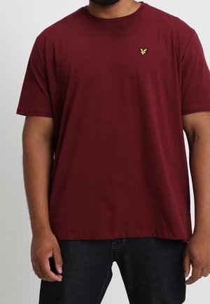 Mężczyzna ubrany w jednokolorowy ciemnoczerwony t-shirt z krótkim rękawem z małym żółtym logo ptaka na lewej piersi oraz ciemnoniebieskie dżinsy.