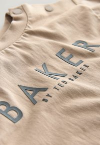 Béžová látka s vroubkovanou strukturou, na které jsou vyražené nápisy "BAKER" a "by TED BAKER" v tmavě šedé barvě. Dva knoflíkové akcenty na výstřihu.