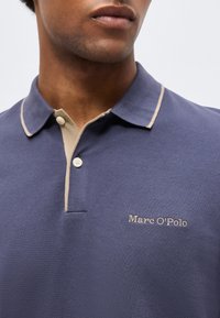 Muž v modrom polokošeli s béžovým lemovaním a gombíkmi, vyšívané logo "Marc O'Polo" na hrudi, detail na hornú časť trupu a krk.