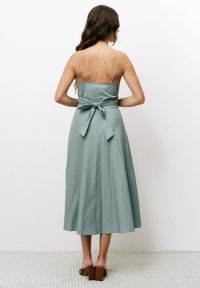 Robe midi verte claire avec des bretelles fines, en tissu texturé et un nœud noué dans le dos. Associée à des chaussures à talons épais marron.