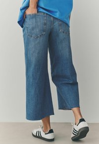 Pantalones culotte de mezclilla en azul medio, con dobladillo sin acabar, dos bolsillos traseros; combinados con zapatos blancos con rayas negras y una blusa azul.