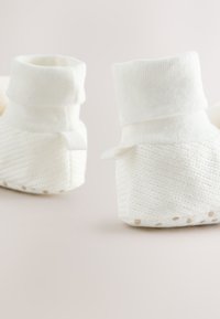 Chaussons blancs en tricot avec un large revers côtelé, corps tissé texturé et semelle en caoutchouc pointillée pour une meilleure adhérence. Agrémentés de petites languettes en tissu.