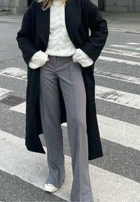 Schwarzer langer Mantel, weißer grob gestrickter Pullover, graue Schlaghose und weiße Sneakers. Das Outfit beinhaltet kuschelige Manschetten und einen lockeren Schnitt.